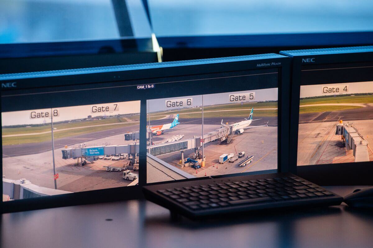 Three monitors display gates at an airport. Two aircraft are parked at the gates and the airport’s runway is visible behind them. 
